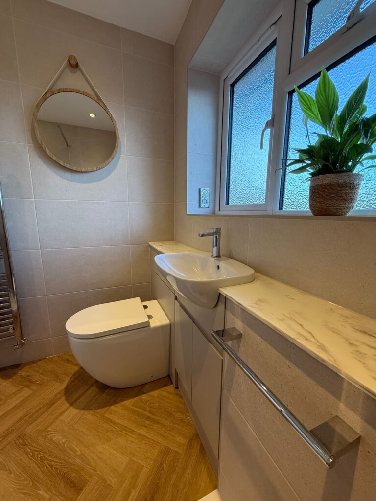 Modern bathroom with beige tiled walls, a round mirror, a white toilet, a sink with a marble countertop, a towel rail, a frosted window, and a potted plant on the windowsill. Brown wood-patterned floor.