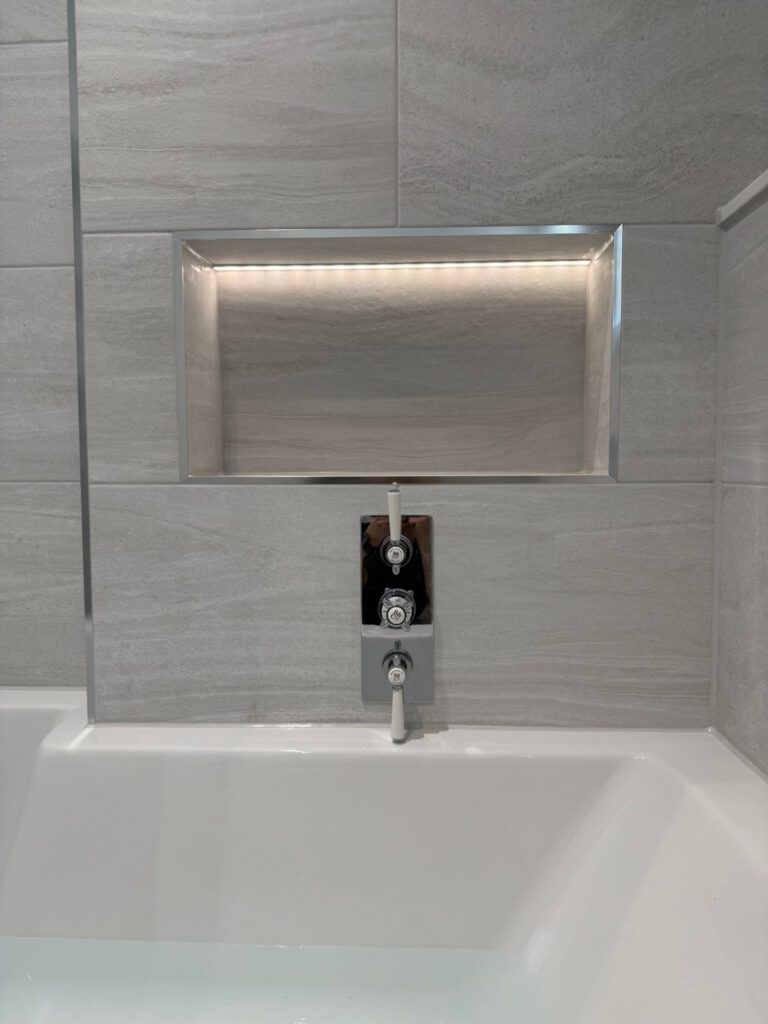 A modern bathtub with grey tiled walls, a built-in rectangular niche with LED lighting above the faucet, and chrome bath controls centered below the niche. The tub is partially filled with water.