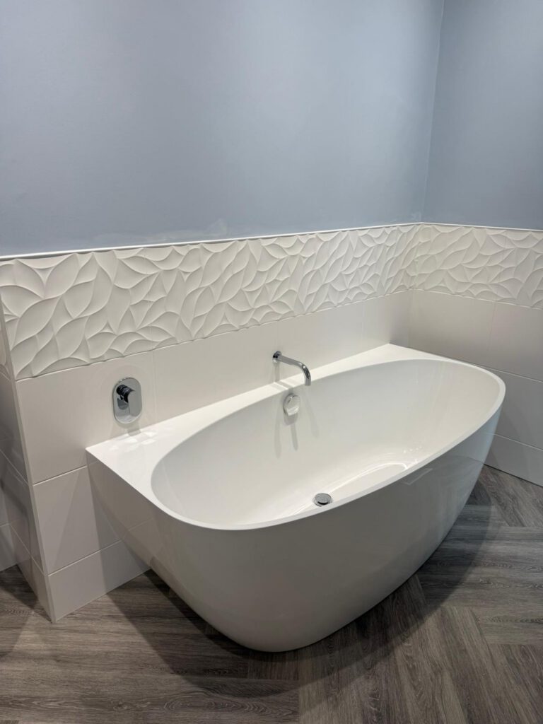 A modern white freestanding bathtub sits against a wall with decorative white textured tiles and a light gray painted upper section, on gray wood-look flooring.