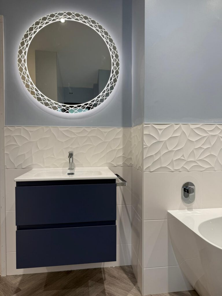 Modern bathroom with a round, backlit mirror above a navy blue floating vanity, white sink, silver faucet, textured white wall tiles, and part of a white bathtub visible on the right.