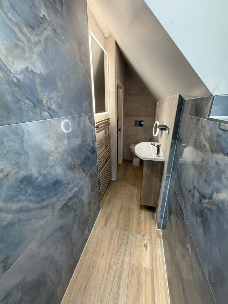 A narrow modern bathroom with large gray marble tiles on the walls, wood-style flooring, a wall-mounted toilet, a sink with a backlit mirror, and a sloped ceiling.