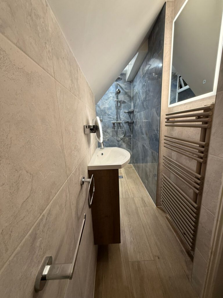 A narrow modern bathroom with beige tile walls, a wall-mounted sink, a large rectangular mirror, a towel warmer, and a walk-in shower with dark gray tiles and a rain showerhead at the far end.