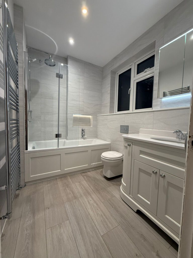 Modern bathroom with light wood flooring, white panel bathtub with glass shower screen, wall-mounted toilet, and a white vanity with sink and mirror. Walls are tiled in light grey and two ceiling lights are on.
