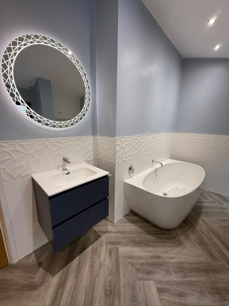 A modern bathroom with a round backlit mirror above a navy floating sink cabinet, a white oval bathtub, textured white wall tiles, and herringbone-patterned wood-look flooring.
