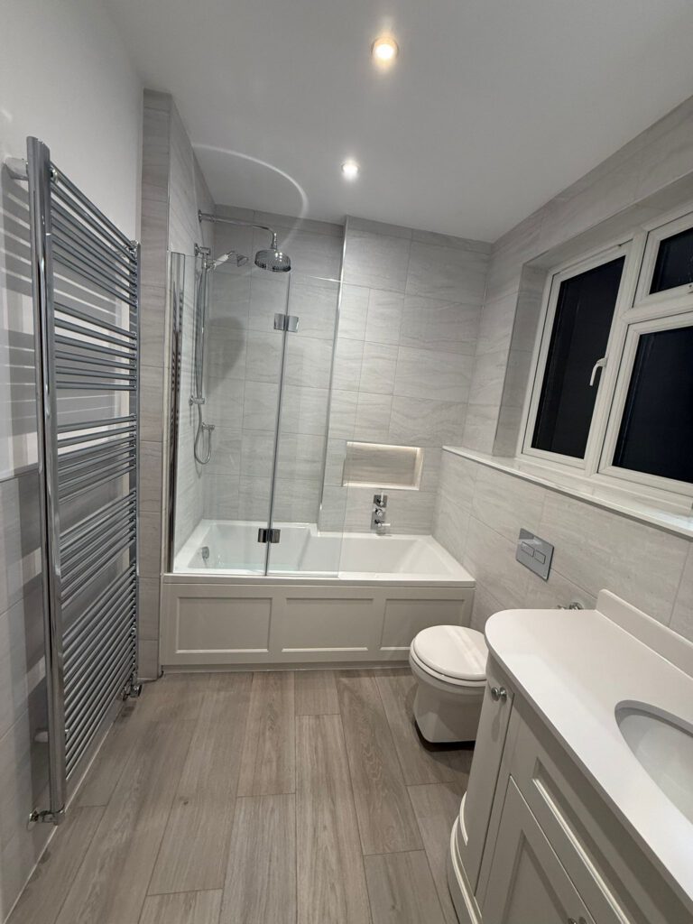 Modern bathroom with light gray tiled walls and floor, a bathtub with a glass shower screen and overhead shower, a white toilet, a white sink with a curved vanity, a heated towel rail, and two windows.