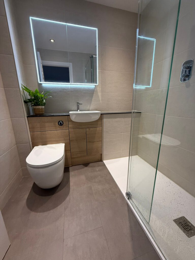 Modern bathroom with beige tiles, a wall-mounted toilet, a small sink with a wooden cabinet, a potted plant, an illuminated mirror, and a glass shower enclosure with a white shower tray.