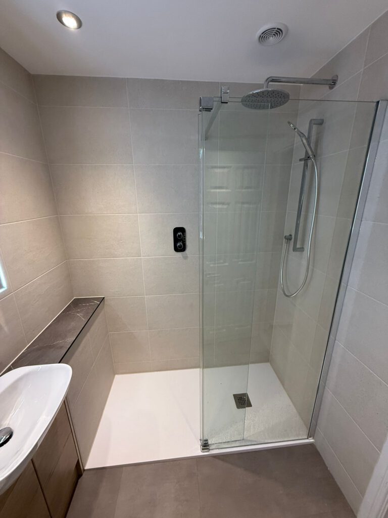 A modern bathroom with beige tiled walls, a walk-in glass shower with a rainfall showerhead and handheld attachment, a wall-mounted black control panel, and a white sink with wooden cabinetry.