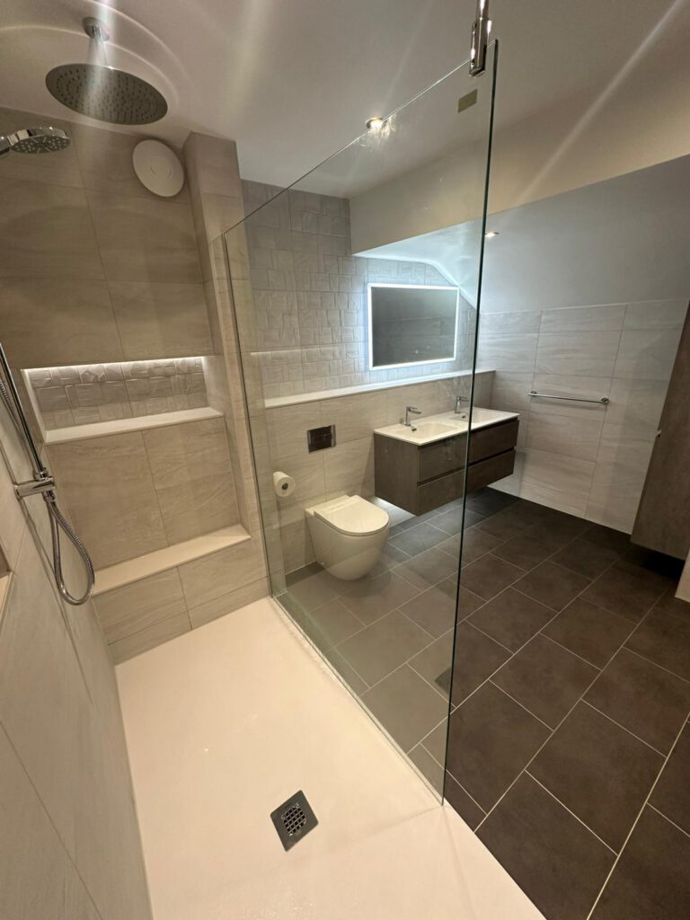 Modern bathroom with walk-in shower, glass divider, wall-mounted toilet, large mirror above a sink, floating vanity, and dark floor tiles. The shower area has built-in shelves and a rainfall showerhead.